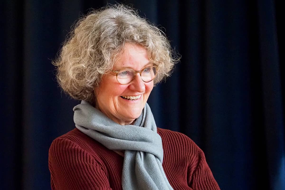 Ein Portraitfoto von Petra Camilla Koch mit lockigem Haar, sie trägt einen roten Pullover und einen hellgrauen Schal. Der Hintergrund besteht aus dunklen, vermutlich schwarzen Vorhängen.